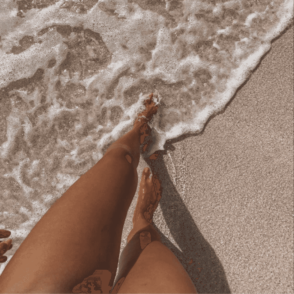 A selfie of a tan womans legs as she walks through the surf at the beach